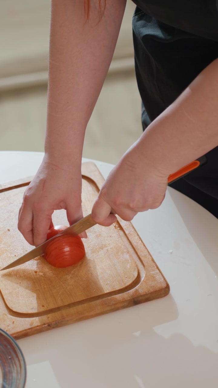 Mani adulte preparano un pomodoro maturo su un tagliere di legno, ripreso dall'alto, con un coltello che si muove verso il basso attraverso il frutto mentre l'altra mano lo tiene fermo, catturando la preparazione fresca in cucina e il controllo attento.