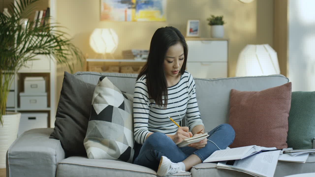 Young Woman In Striped Shirt And Wearing Headphone Studying With Textbooks And Taking Notes On Notebook