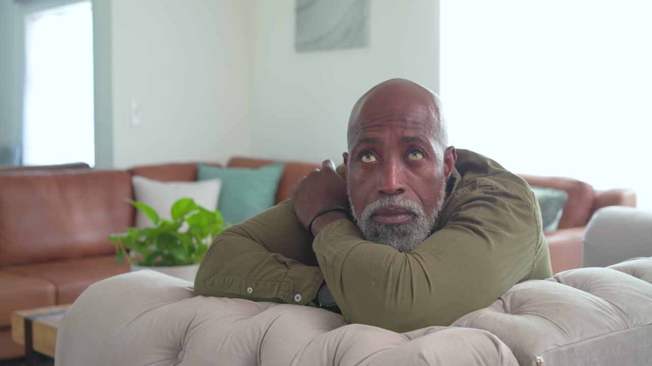 Uomo afroamericano appoggiato a un divano trapuntato beige a casa, che vede la luce della finestra, cambia lo sguardo e sorride