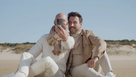 Vorderansicht-Eines-Glücklichen-Schwulen-Paares,-Das-Beim-Sitzen-Am-Strand-Ein-Selfie-Am-Telefon-Macht