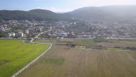 A-small-town-with-the-same-brown-houses-and-roofs,-through-which-a-wide-asphalt-road-passes,-is-located-between-green-fields-and-mountains,-near-some-houses-there-is-a-pool-with-clear-blue-water