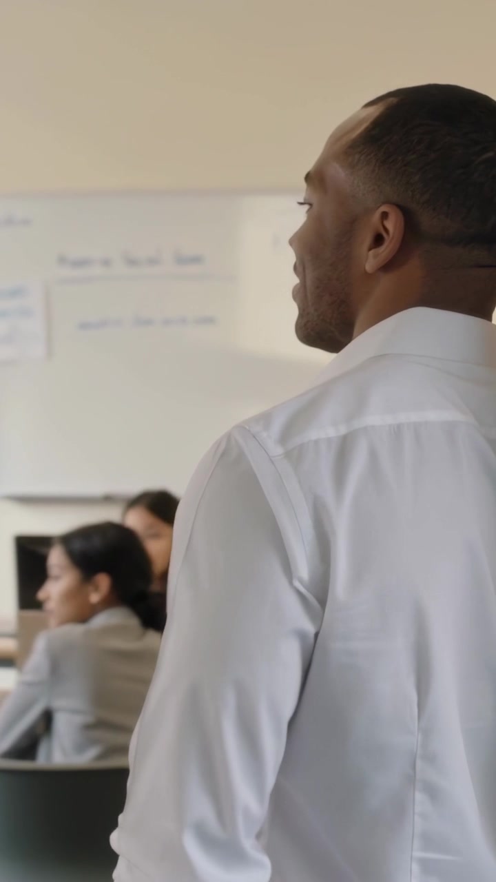 Vertical video: Entering teacher in white shirt walking in classroom, pausing, teaching with laptop