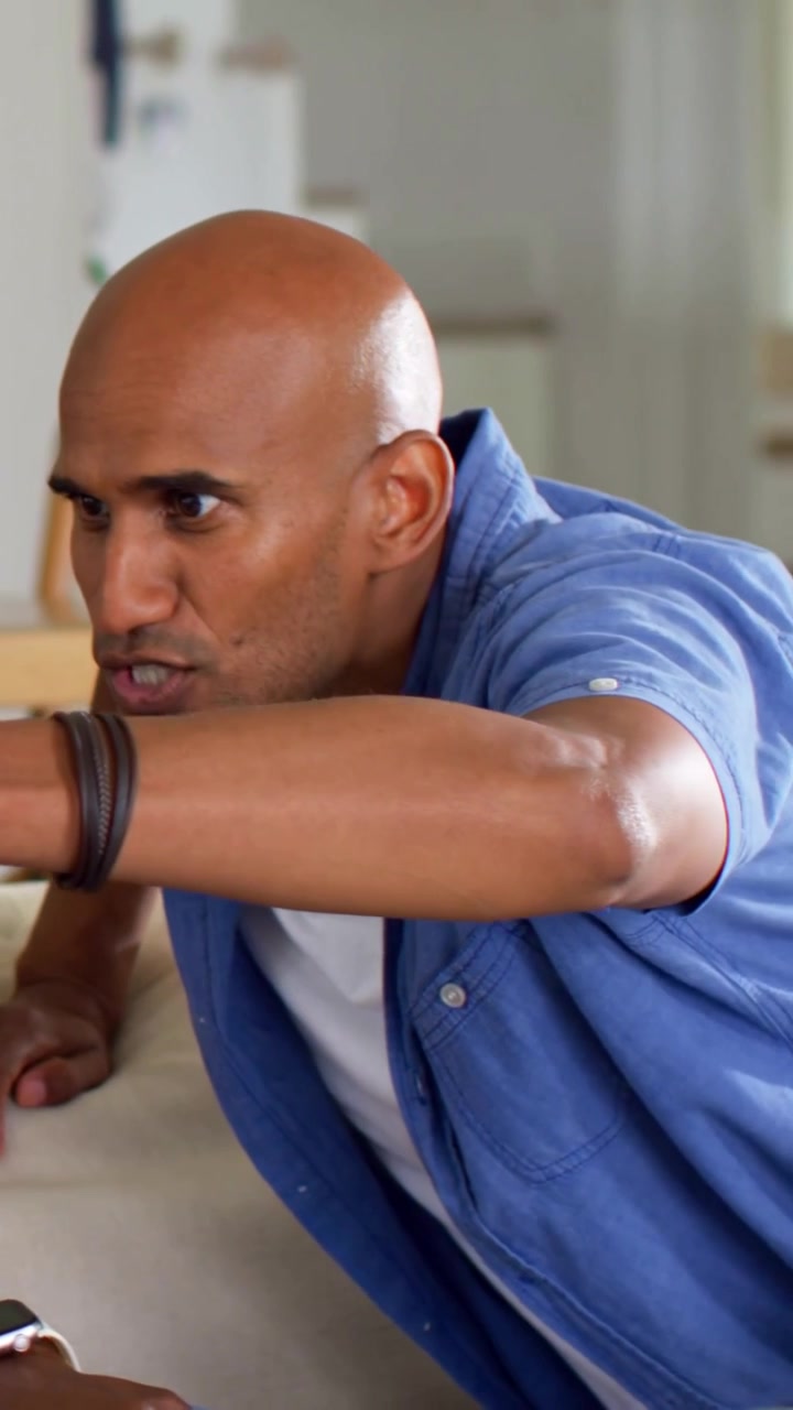 Vertical video: Leaning blue shirt man reacting to offscreen cue, talking on couch with green beers