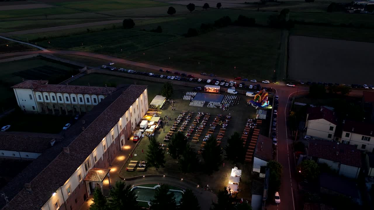 Static drone view at night of the Festa di San Bernardo beside Chiaravalle della Colomba Abbey, with glowing tents, dining tables, and crowds gathered around the illuminated monastery