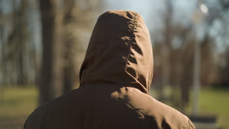 a close- up of an individual wearing a brown hooded jacket walking outdoors, with a blurred background of trees and a pathway