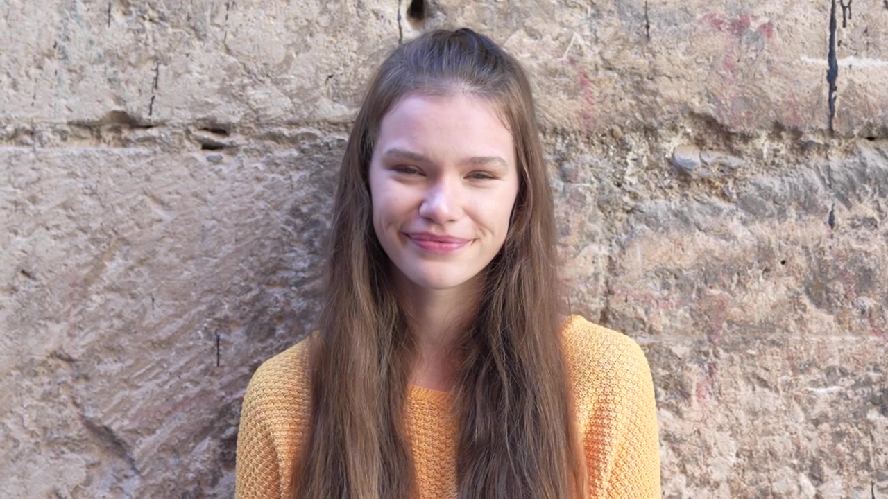 Una giovane donna sorridente che indossa un maglione accogliente posa contro un affascinante muro di pietra.