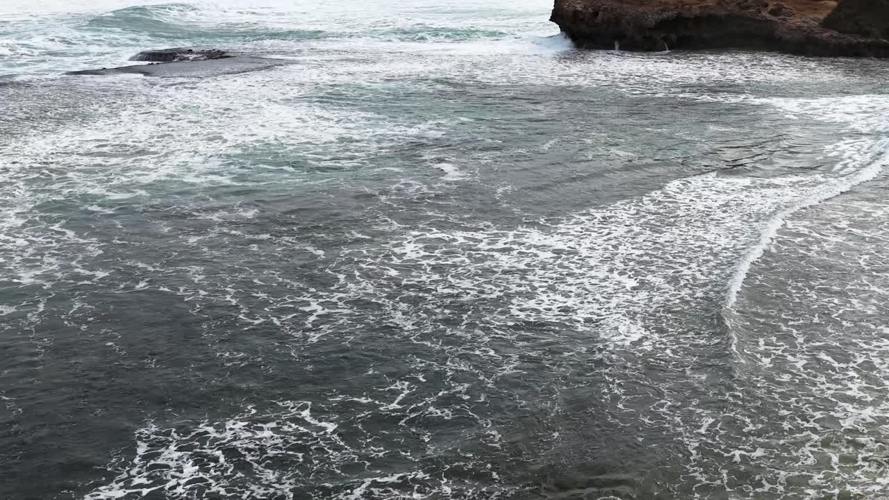 Drone captures waves rolling onto sandy beach and rocky cliffs under overcast natural daylight