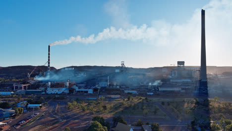 Aerial,-Track-right,-Industrial-smoke-billowing-from-chimney-stacks-from-the-Mount-Isa-mines-against-blue-sky-background,-Mount-Isa