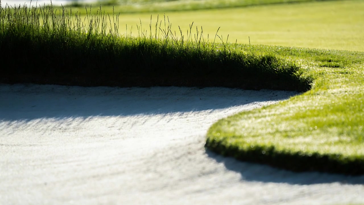 Liikkuva kamera paljastaa golfkentän kaarevan bunkkerin reunan, korostaen haravoitua hiekkaa ja nurmea.