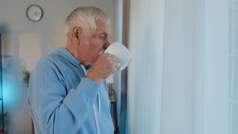 Smiling-senior-old-grandfather-man-drinking-a-cup-of-coffee-or-herbal-tea-at-home-room-near-window