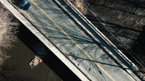 drone ascends above calm empty bridge as sedan and suv drive across asphalt, long tree shadows