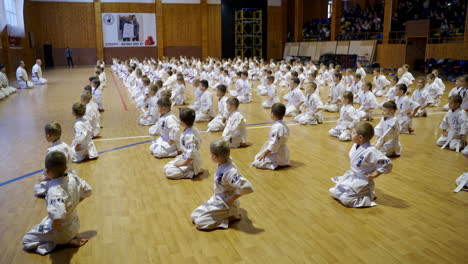 large gym with numerous kids sitting on their knees on the floor. children bend to greet the masters. spectators sitting at the backdrop.