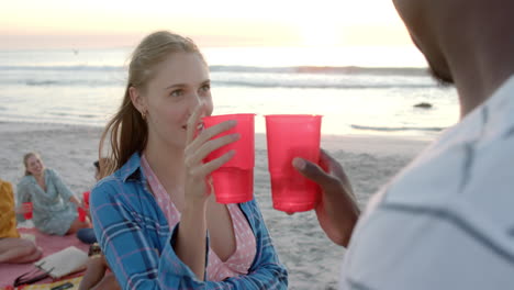 joven mujer caucásica beba de una taza roja en una fiesta de playa, frente a un hombre afroamericano