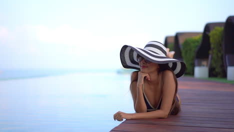 Elegante-Mujer-Exótica-Acostada-Junto-A-La-Piscina-En-Traje-De-Baño,-Gafas-De-Sol-Y-Sombrero-Disfrutando-Y-Relajándose-En-Sus-Vacaciones-De-Verano,-Fotograma-Completo