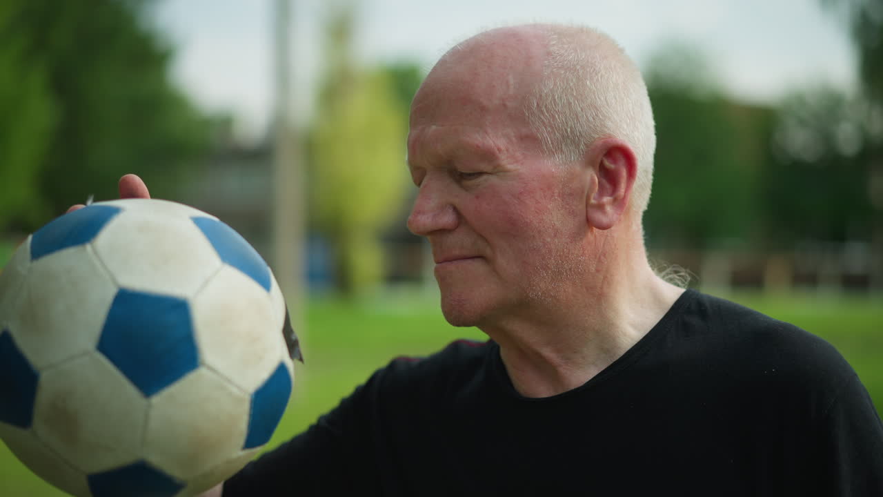 An elderly man in a black top is thoughtfully observing a soccer ball held close to his face, with a blur view of building in the background