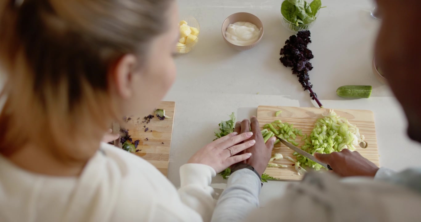 Couple chopping boards with knives at counter, ingredients set, woman guiding hand making salad