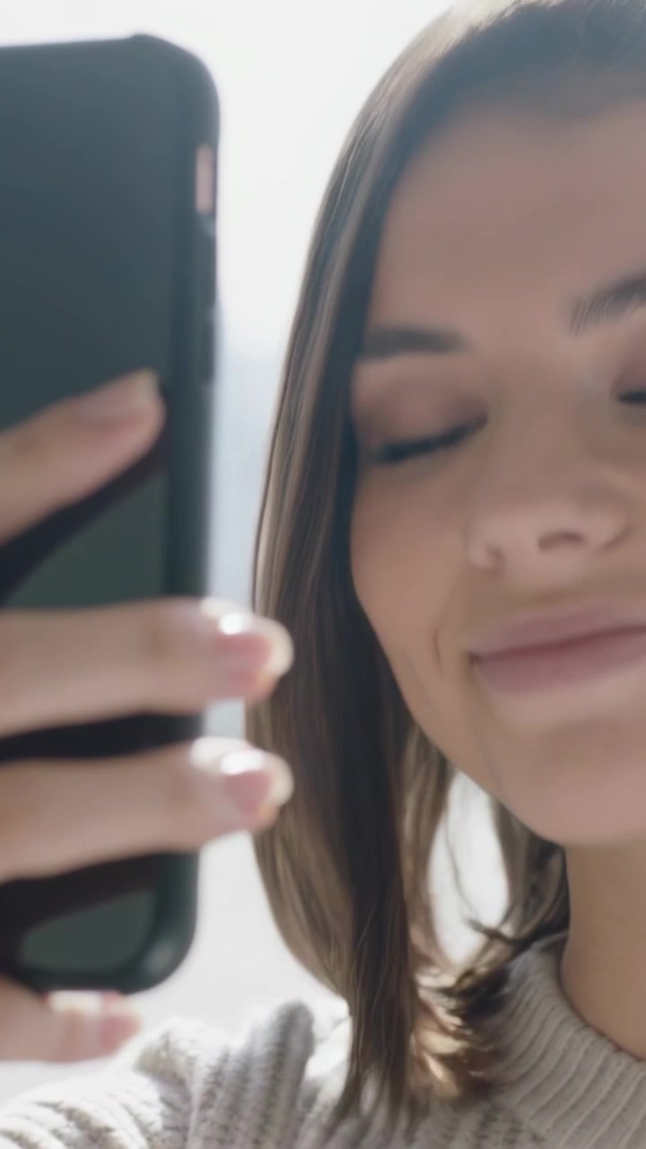 Video verticale: Donna che tiene un cellulare, in posa vicino a una finestra, che tocca lo schermo, con top in maglia, mani, spazio per il testo