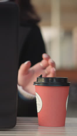 vista trasera de primer plano de la computadora portátil en la mesa, persona con abrigo negro sentada con una taza de café dejándola caer en la mesa en un entorno de restaurante, fondo borroso