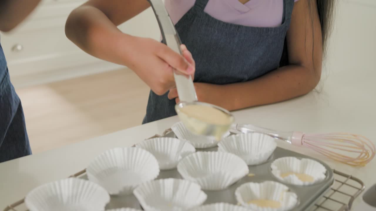 Afroamerikansk mor veileder et barn iført forkle, som øser røre med en øse for å bake cupcakes på kjøkkenet.
