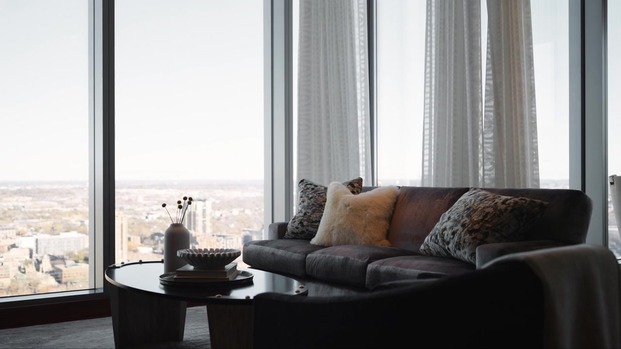 Soggiorno elegante con un divano sontuoso, cuscini decorativi e grandi finestre che offrono una vista panoramica sulla città, creando un'atmosfera accogliente e raffinata in un grattacielo di lusso.