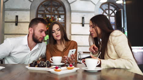 a group of men and women in office clothes sitting in a cafe at a large table and show each other in mobile phones funny pictures