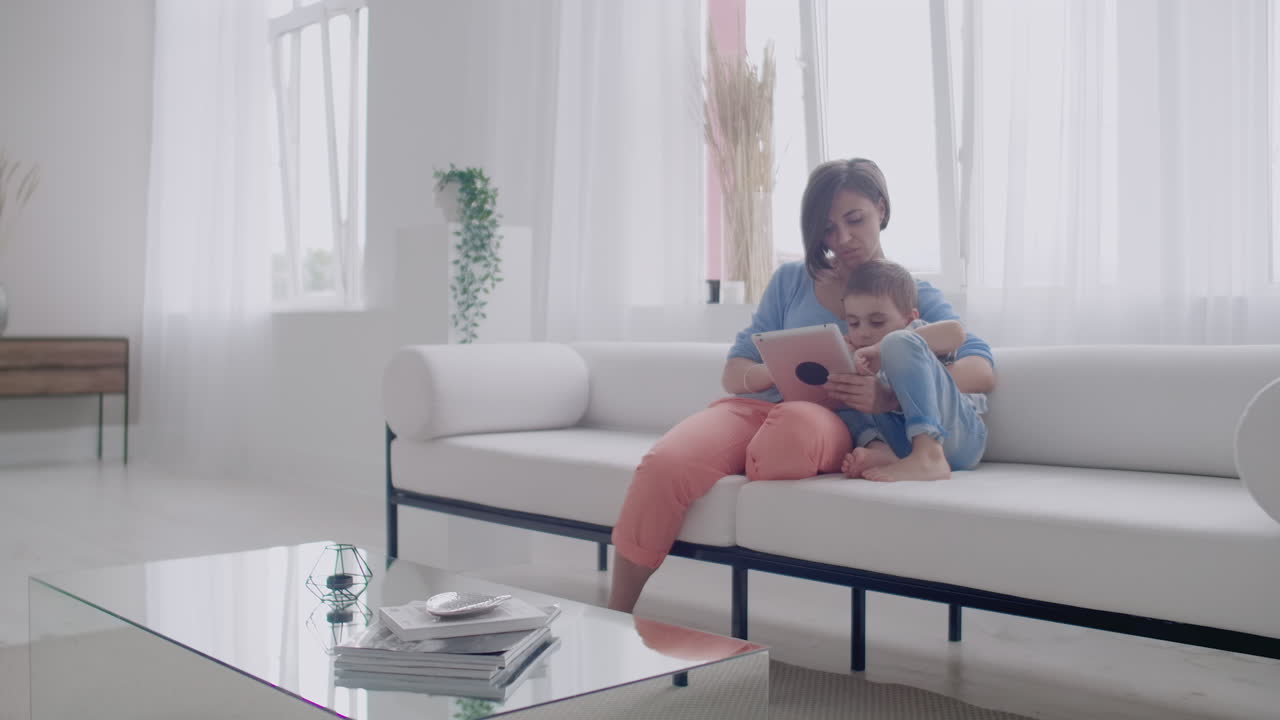디지털 태블릿을 사용하여 소파에 앉아있는 엄마와 아들이 함께 비디오를 보는 터치 스크린을 가진 태블을 사용하는 행복한 엄마와 작은 소년. 웃는 엄마와 귀여운 소년은 디지털 태블에서 놀고 있습니다.