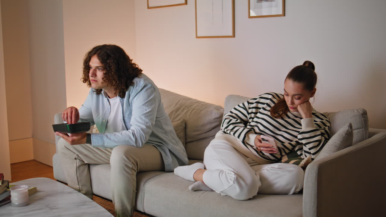 Ignoring couple sitting home sofa. Upset woman looking smartphone husband eating