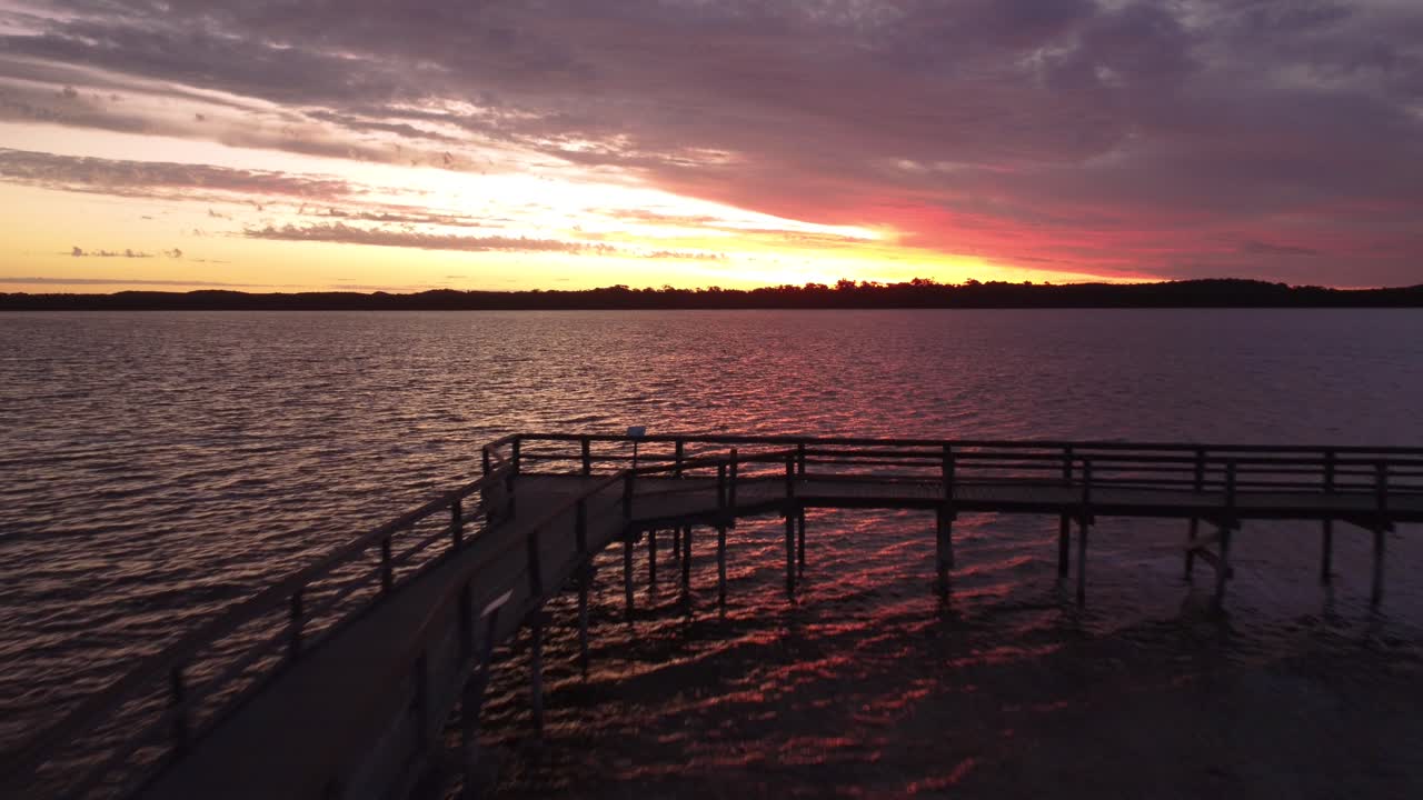 Riprese aeree con drone che si allontanano all'indietro da un pontile di legno su una riva di lago ghiacciato durante uno splendido tramonto invernale, rivelando il paesaggio scenografico con nuvole colorate e acqua calma