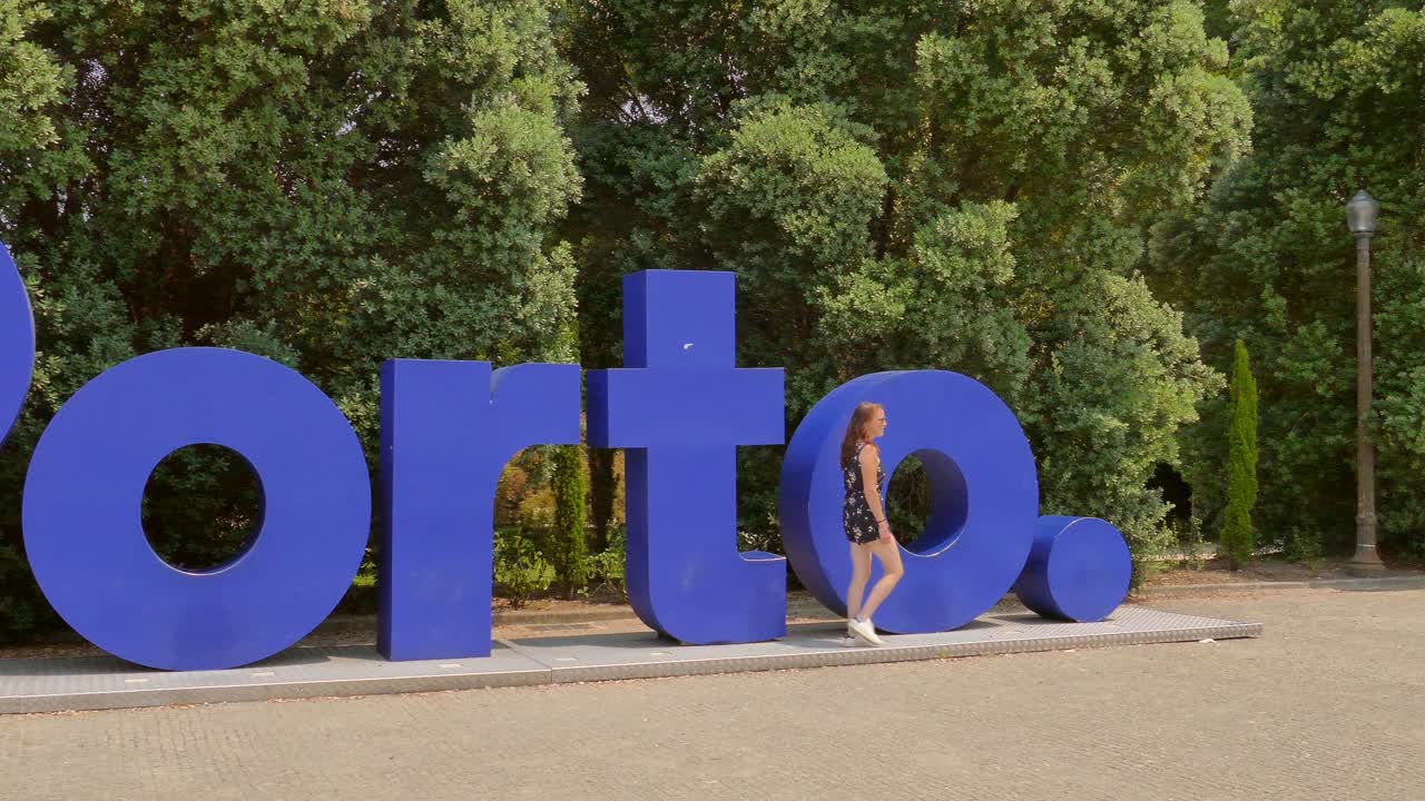 Young tourist woman walking left to right in front of a large blue sign reading &amp;quot;Porto&amp;quot;, establishing the cities name