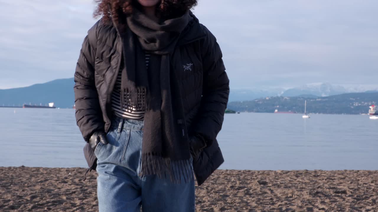 Youg woman wearing black beanie, black puffy coat and jeans on a beach walking towards the camera on a beautiful winter day with boats and oil tankers in the background (4K)