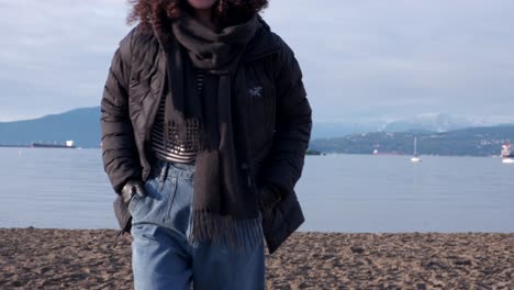 youg woman wearing black beanie, black puffy coat and jeans on a beach walking towards the camera on a beautiful winter day with boats and oil tankers in the background (4k)