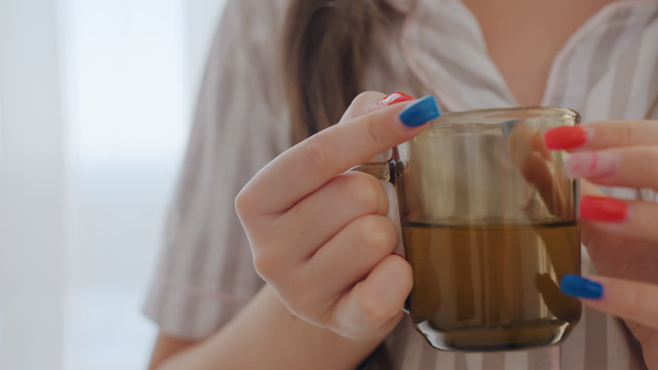 Donna tranquilla con unghie vivaci, primo piano di una donna che tiene una tazza durante una serena pausa mattutina, scena rilassante che mostra una donna con unghie colorate che sorseggia il tè vicino alla finestra di casa alla luce del mattino