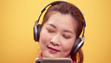 밝은 노란색 배경에서 편안한 헤드폰으로 음악을 즐겁게 듣기 위해 디지털 태블릿과 스트리밍 응용 프로그램을 사용하여 아시아의 멋진 젊은 여성을 닫습니다.