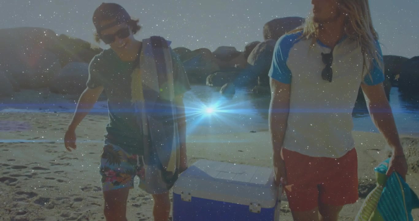 Due uomini che trasportano un frigo portatile blu, camminando verso una caletta per il tempo libero in spiaggia, con il sole in controluce che crea un effetto a stella