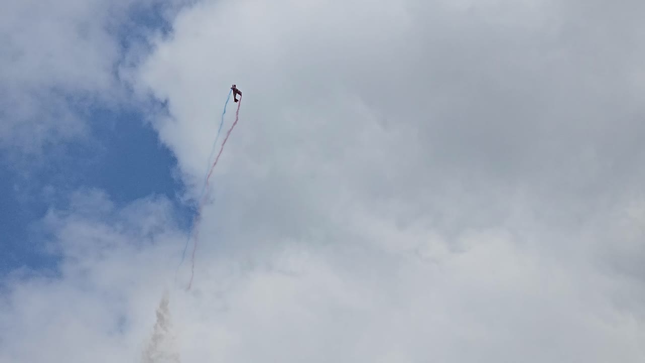 A stunt plane performs a dramatic aerial trick, leaving red, white, and blue smoke trails against a cloudy sky in a dynamic display.