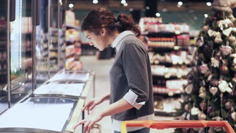 Young-beautiful-brunette-girl-comes-up-to-a-freezer-trying-to-decide-which-product-to-buy-looking-at-price-tag.-Shopping-at-grocery-store