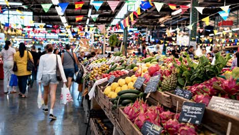 shoppers are browsing through a lively market aisle filled with colorful fresh fruits, vegetables, and international flags hanging overhead, creating a vibrant and multicultural atmosphere