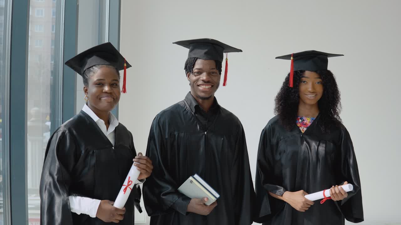 Three African-American university graduates with diplomas and books in their hands stand by a window with a gray frame. Students are dressed in a festive robe and a master's hat