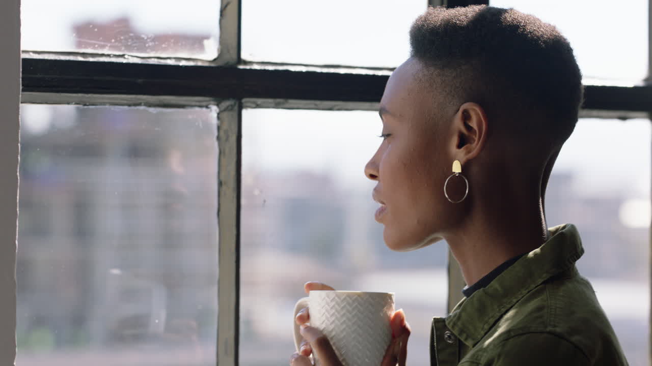 felice donna afroamericana che beve caffè a casa godendosi l'aroma guardando fuori dalla finestra pianificando in anticipo rilassandosi donna nera sorridente soddisfazione