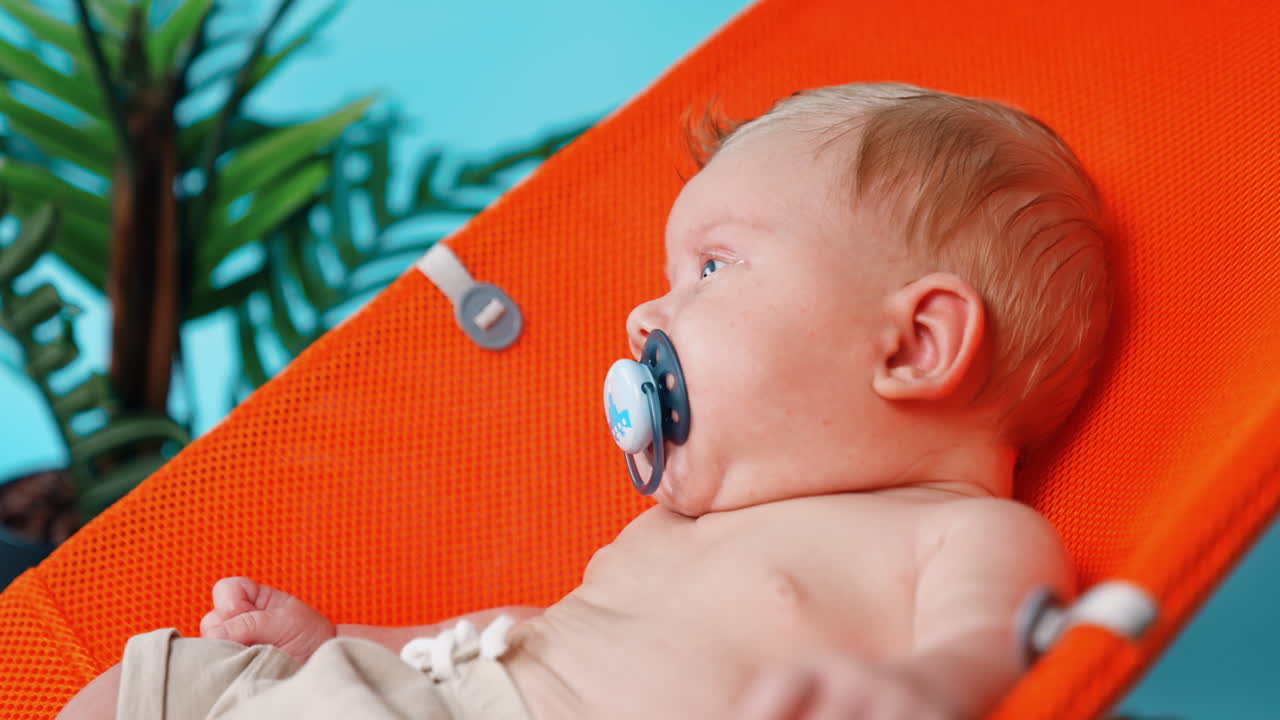 Cute blonde Caucasian infant with pacifier. Baby boy resting in the orange chair. Close up.
