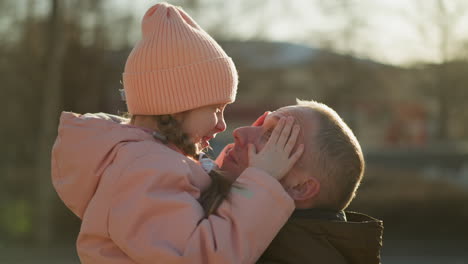 a joyful little girl in a pink cap and jacket is being carried by a man. she is playfully touching her nose to his nose, both smiling warmly and sharing a tender, happy moment outdoors