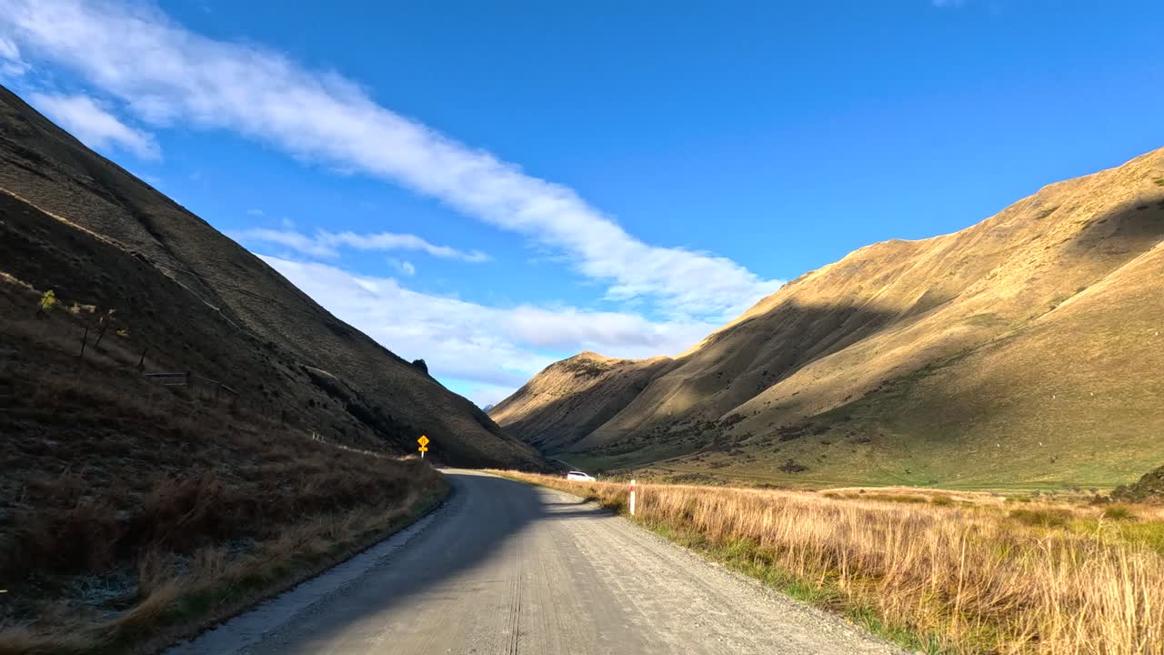 Vehicle travels along gravel road through sunlit mountain valley, wide angle, steady camera, daylight