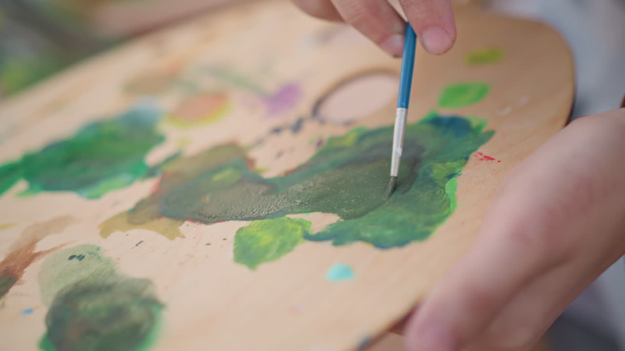 Primo piano del processo di miscelazione della pittura. Immagine dettagliata che cattura le mani mentre mescolano i colori su una superficie di legno. Fotografia intima in studio che mostra vivaci pozze di pigmento sapientemente mescolate