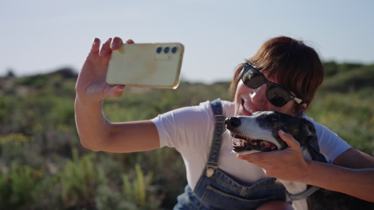 Cena em câmera lenta de mulher sorridente tirando uma selfie com seu cachorro ao ar livre enquanto a câmera se move em um movimento circular suave. Momento de estilo de vida feliz, mídias sociais e companhia de animais de estimação.