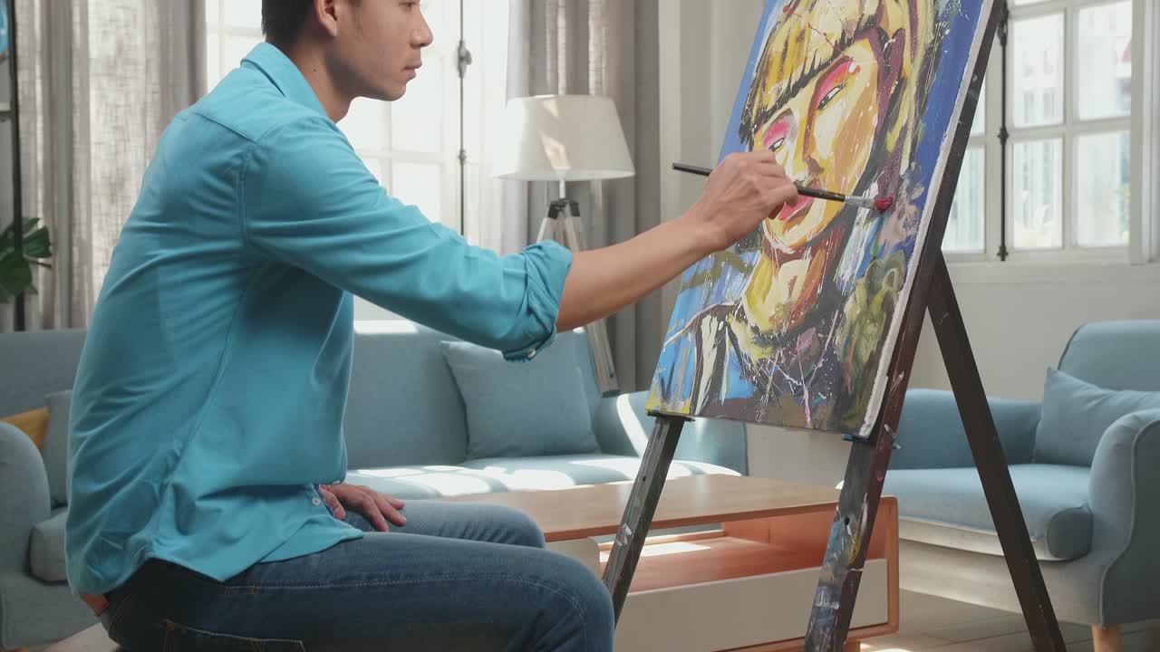 Medium Shot Side View Of An Asian Artist Man Sitting On Round Wooden Chair Without Backrest Holding Paintbrush And Mixing It With Colour Before Painting A Girl'S Hair On The Canvas