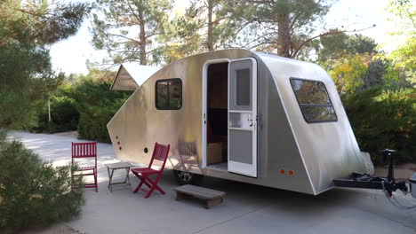 a new deluxe modern teardrop travel trailer with chairs out for people to enjoy nature while camping in the green forest with trees