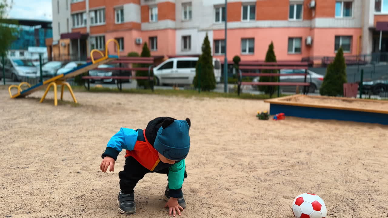Lovely Caucasian baby boy picks up the ball from the ground. Little toddler distracts on the sand and touches it with his hand.