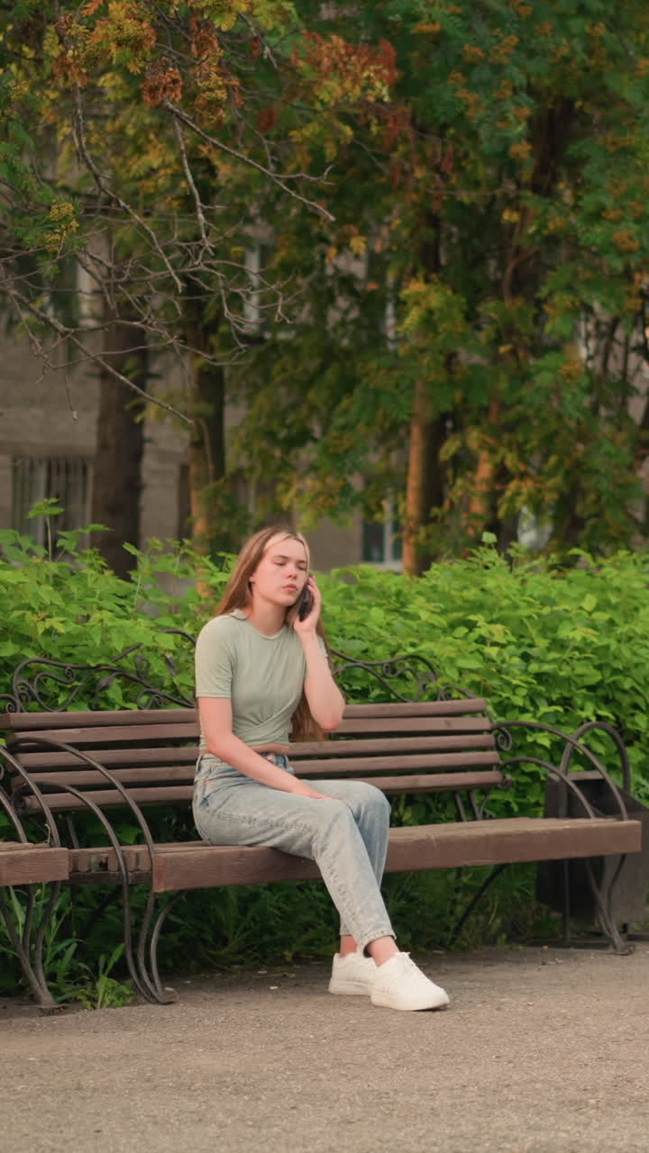 전화 통화 중인 여인은 나무 벤치에 앉아서, 잎자루를 통해 어내는 은 초록색과 나무, 그리고 뒷면에 있는 건물에 둘러싸여 생각 깊게 내려다보고 있습니다.