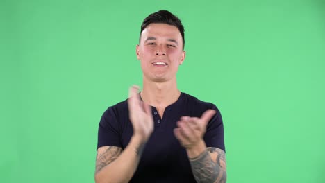 retrato de un joven guapo está mirando directo y aplaudiendo sus manos indiferente. bruneta ardiente con cabello elegante en una camiseta negra y tatuajes en las manos en una pantalla verde en un estudio