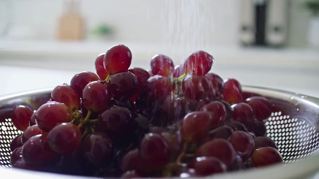 Cena em close-up de ângulo baixo mostra uvas vermelhas dentro de um escorredor de aço inoxidável enquanto um fluxo constante de água corre sobre a fruta, criando respingos, gotículas, brilho e uma atmosfera de preparação de alimentos limpa e fresca.